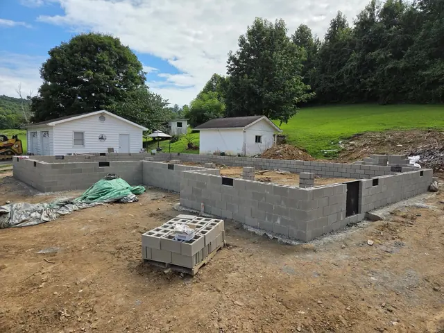 Block foundation walls on a country lot with outbuildings and hardwood trees behind.